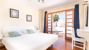Habitación de hotel con una cama grande, un escritorio y un balcón con vistas a palmeras y edificios.