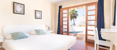 A hotel room with a large bed, a desk, and a balcony with a view of palm trees and buildings.