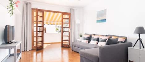 Een woonkamer met een grijze bank, een televisie op een dressoir, een plafondventilator en een uitzicht door een deur op een terras.