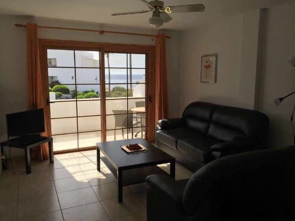 Ein Wohnzimmer mit einer Couch, einem Couchtisch und einem Fernseher. Eine Glastür führt zu einem Außenbereich mit Blick auf das Meer.