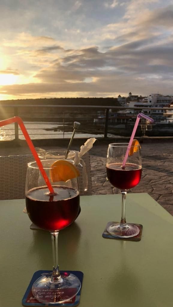 Dos vasos de bebida roja con pajitas y una rodaja de naranja sobre una mesa con vistas a la puesta de sol y una masa de agua.