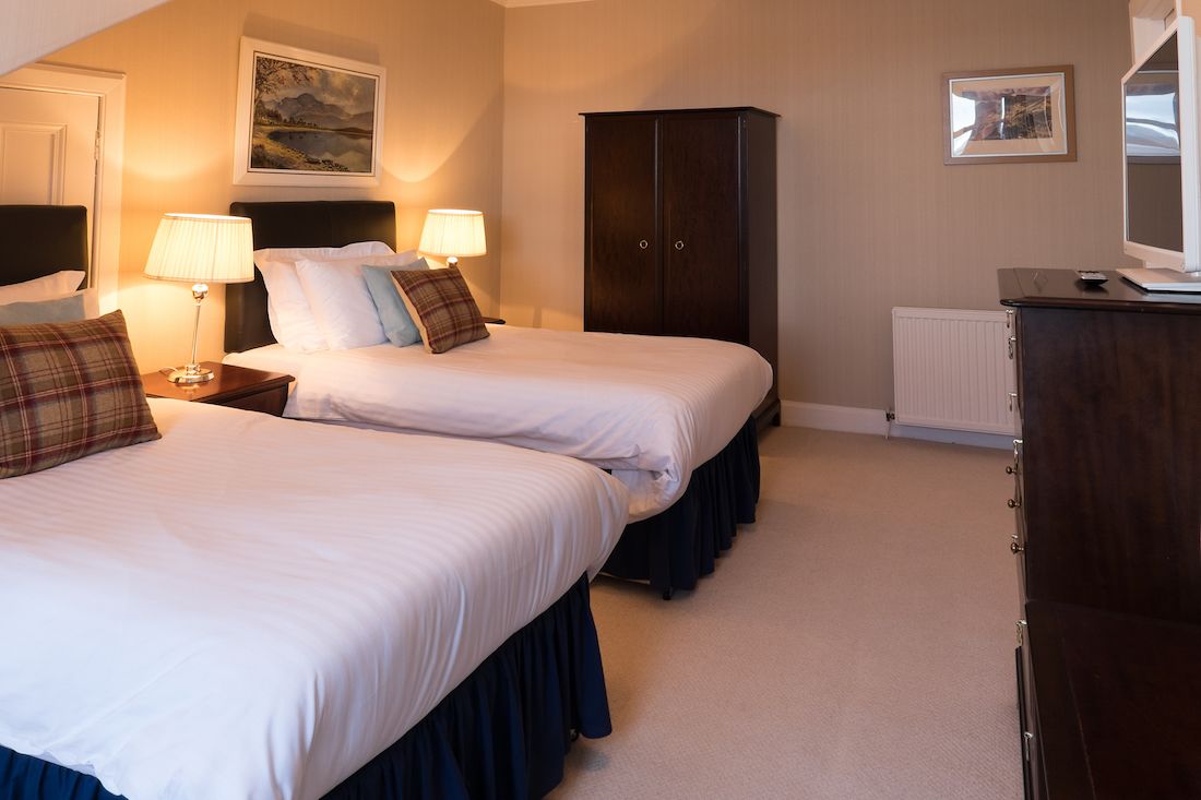 A hotel room with two beds, a wardrobe, a desk with a lamp, and a radiator.