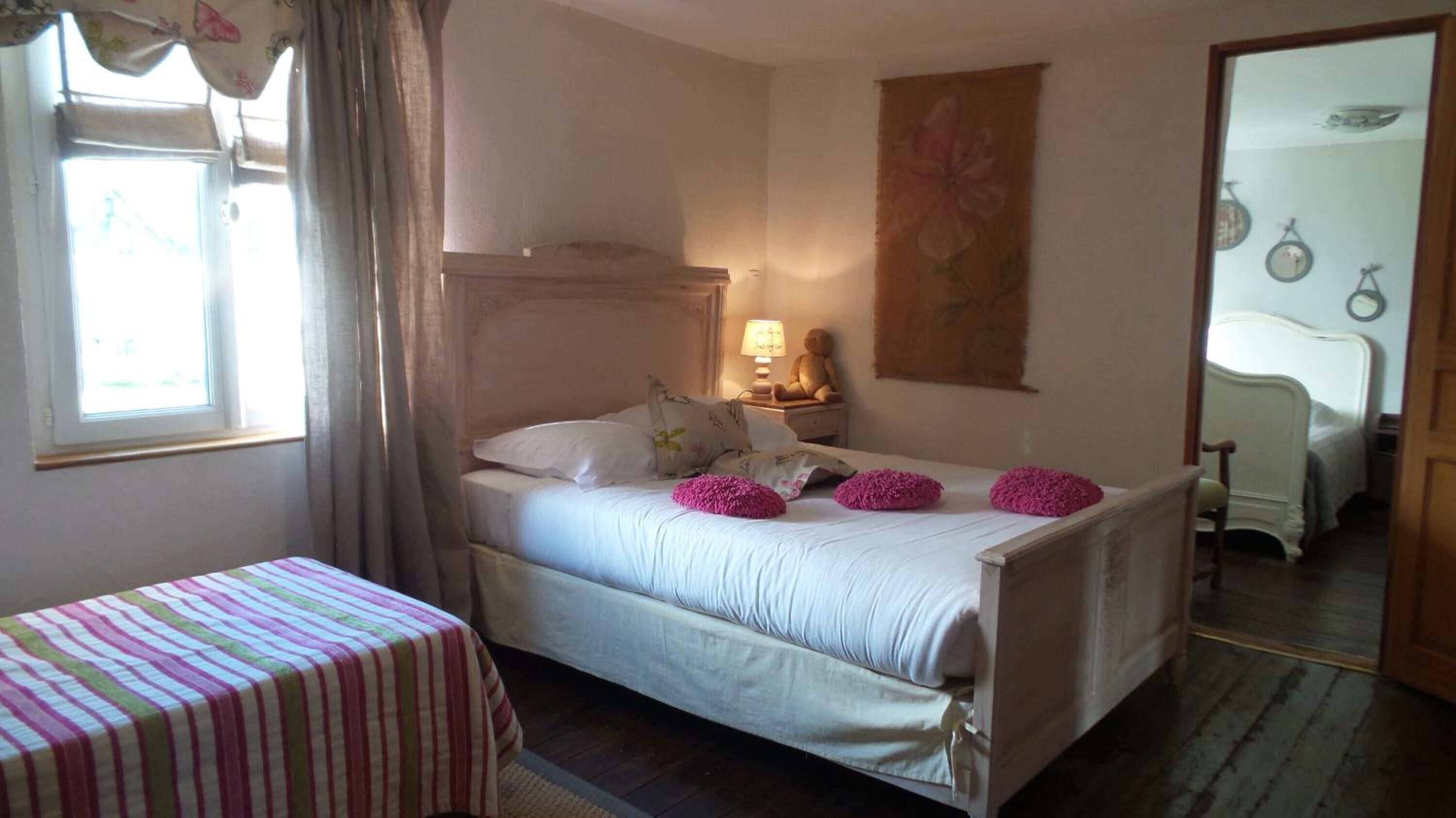 A bedroom with a bed, a bedside table with a lamp, a window with curtains, and a mirror on the wall.