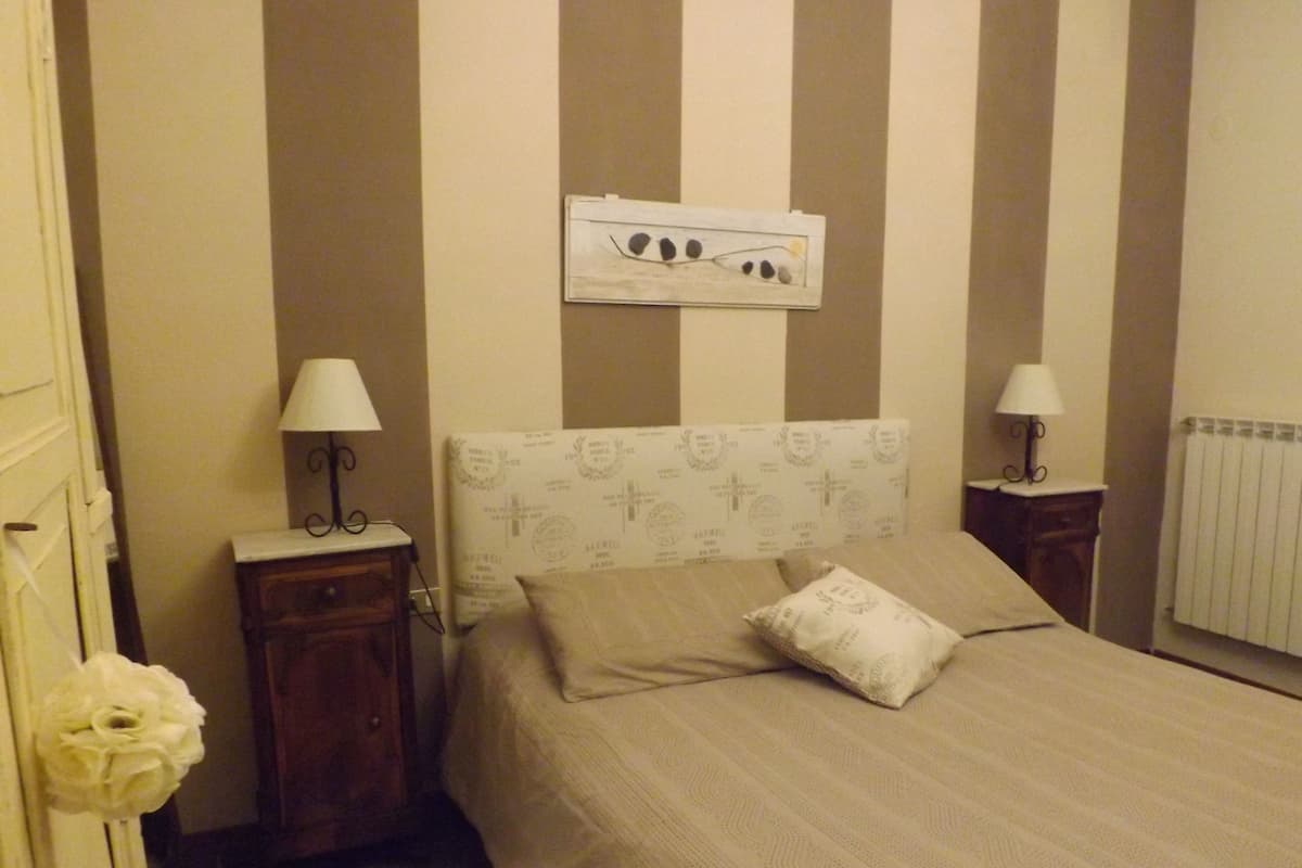 A bedroom with a striped wall, a bed with a pillow, a nightstand with a lamp, and a radiator.