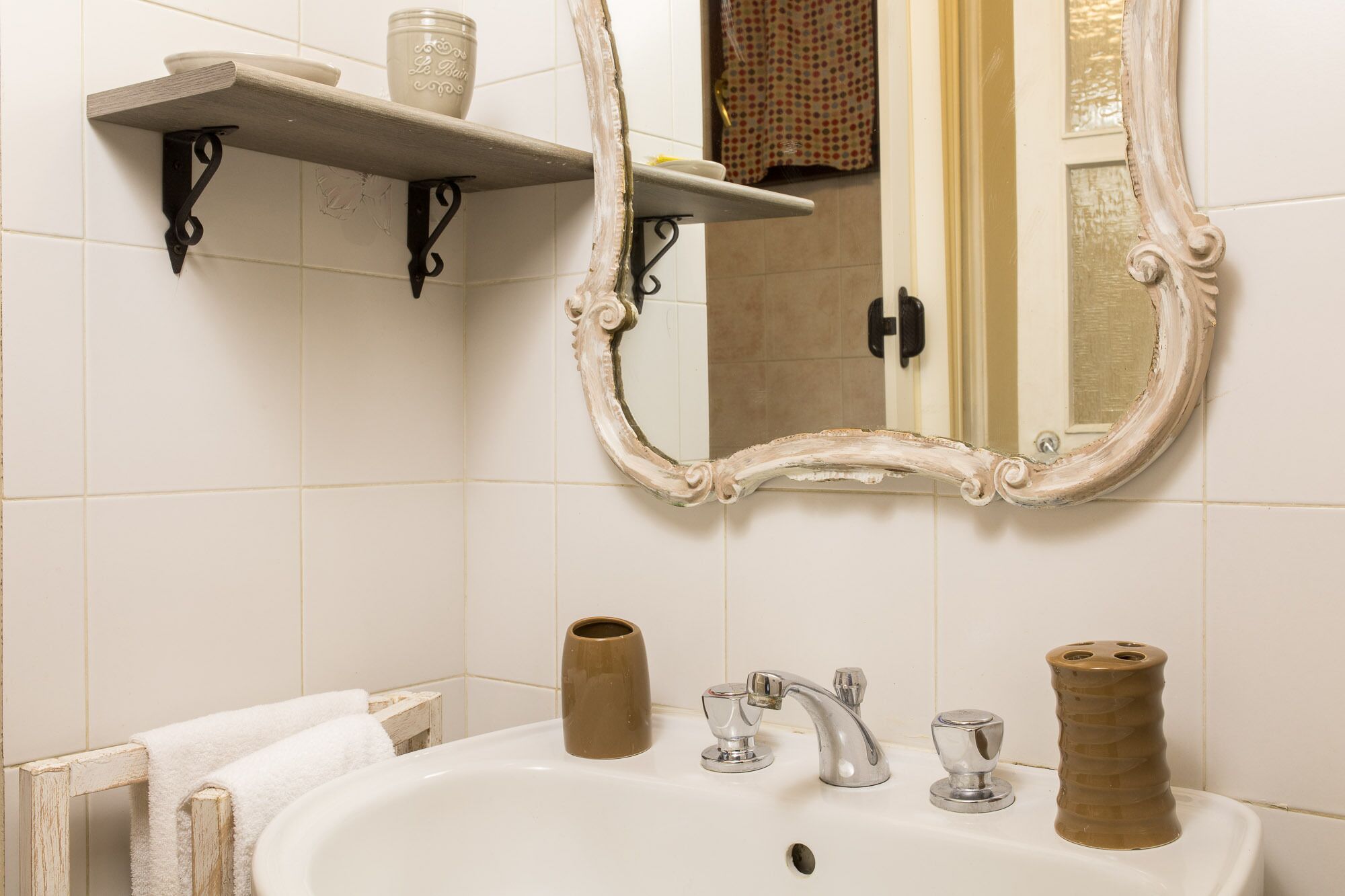 Un bagno con un lavandino bianco, uno specchio con cornice decorata, uno scaffale con un contenitore e un porta asciugamani.