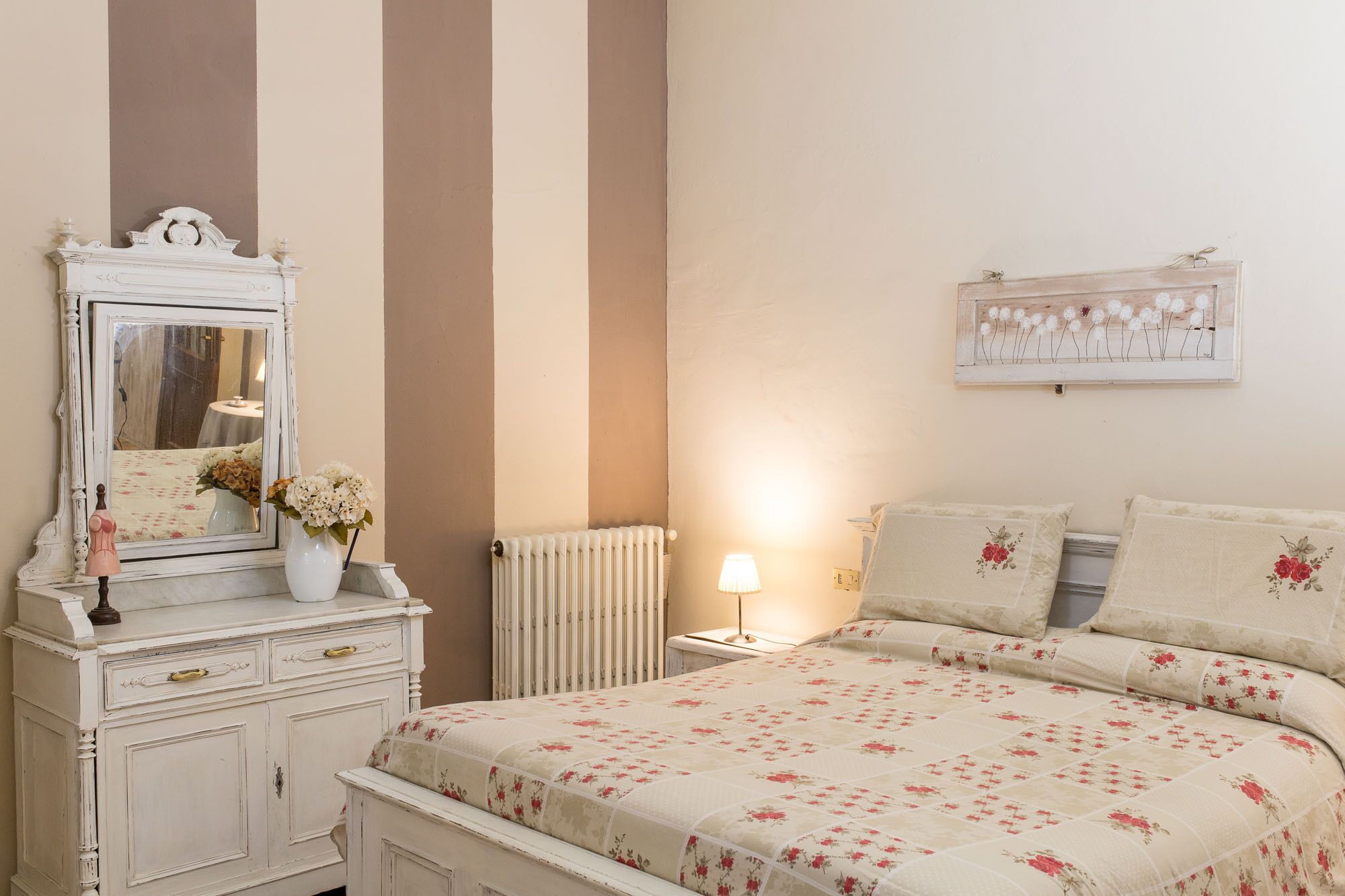 A bedroom with a bed, dresser, mirror, lamp, and floral artwork on the wall.
