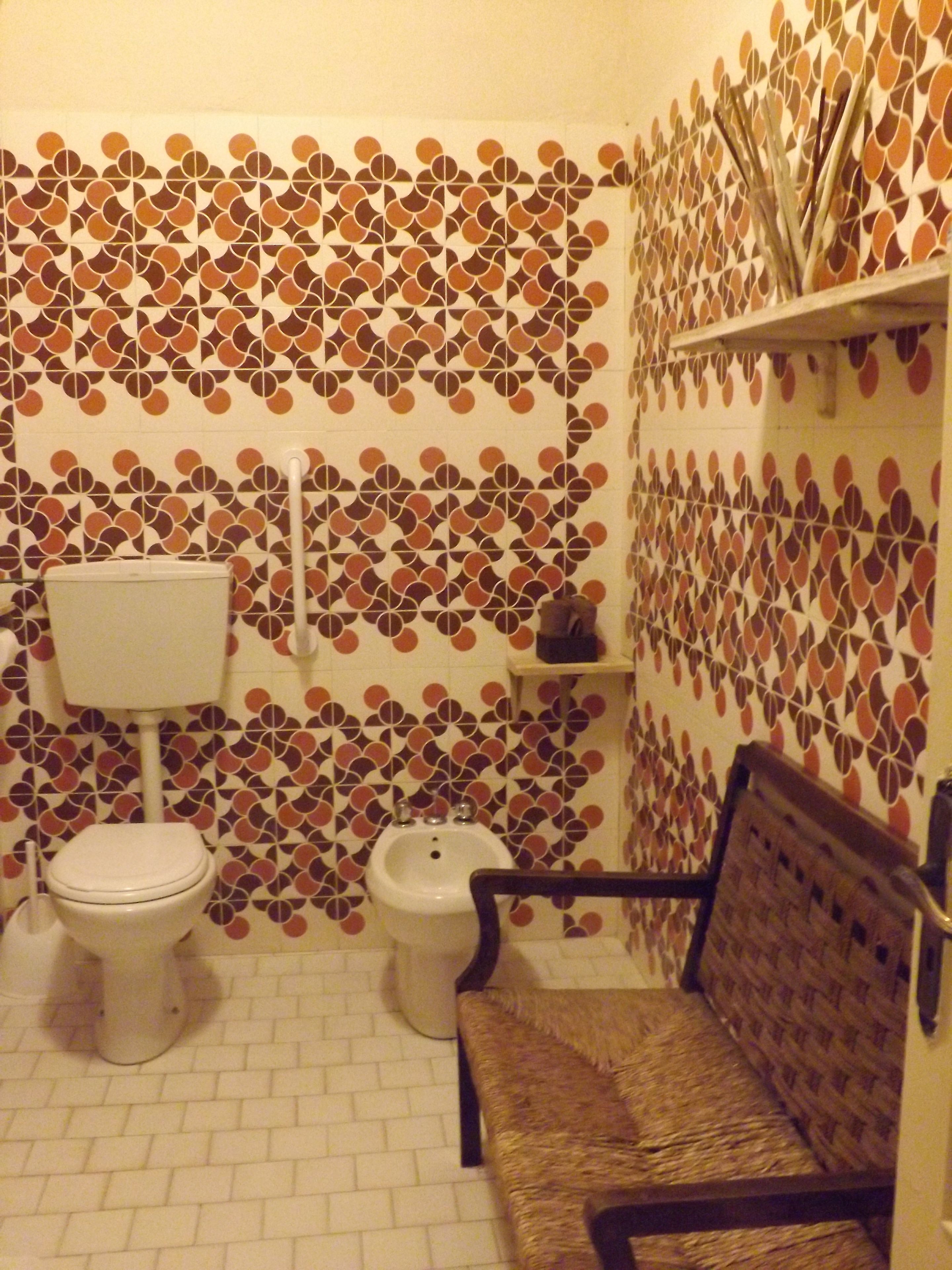 Een badkamer met een betegelde muur met een patroon, een toilet, een bidet en een zitbankje.