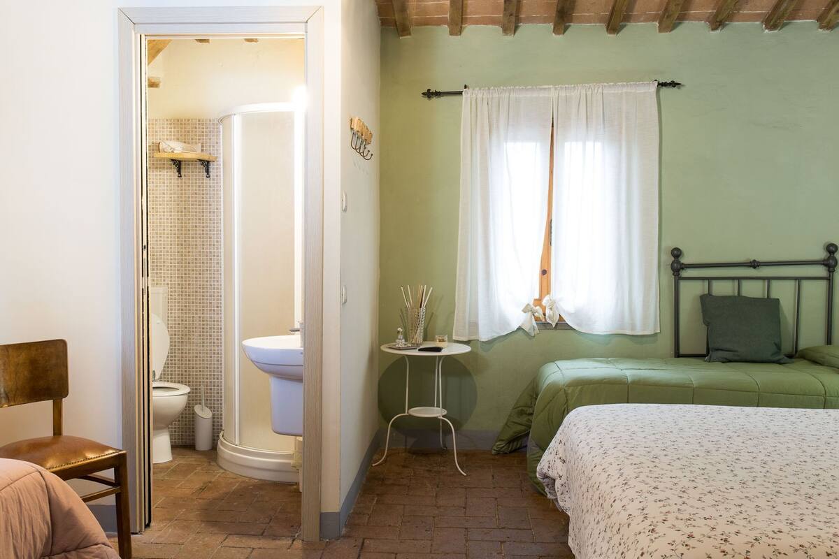 A bedroom with a bed, a window with curtains, a bedside table, and a bathroom with a toilet and shower.