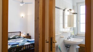 A bedroom with a bed, a bedside table with a lamp, a ceiling fan, and a bathroom with a sink, mirror, and towel rack. - Manzoni Apartment Le Castella (Le Castella)