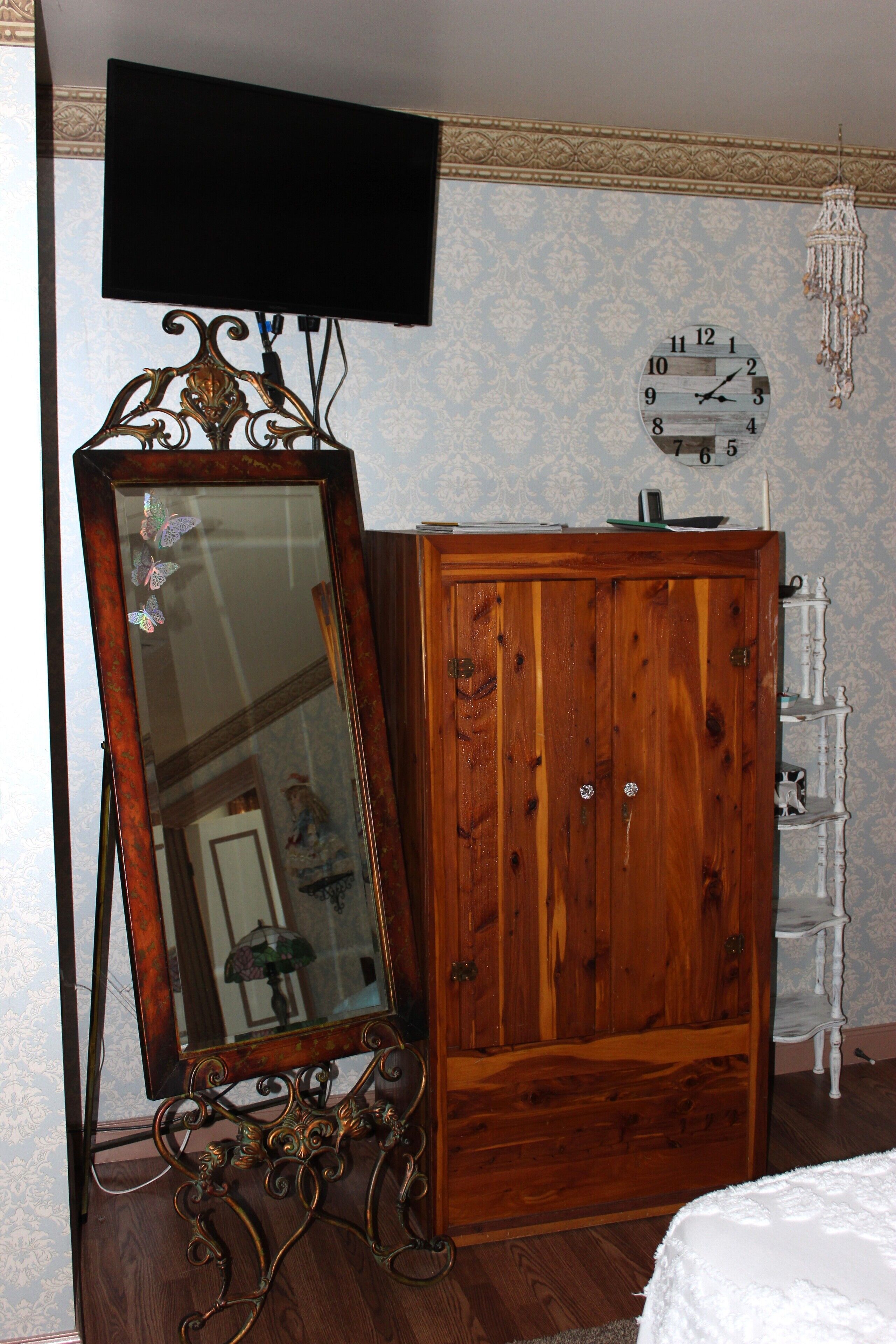 A room with a wooden wardrobe, a large mirror with a decorative frame, a wall clock, and a television mounted on the wall.