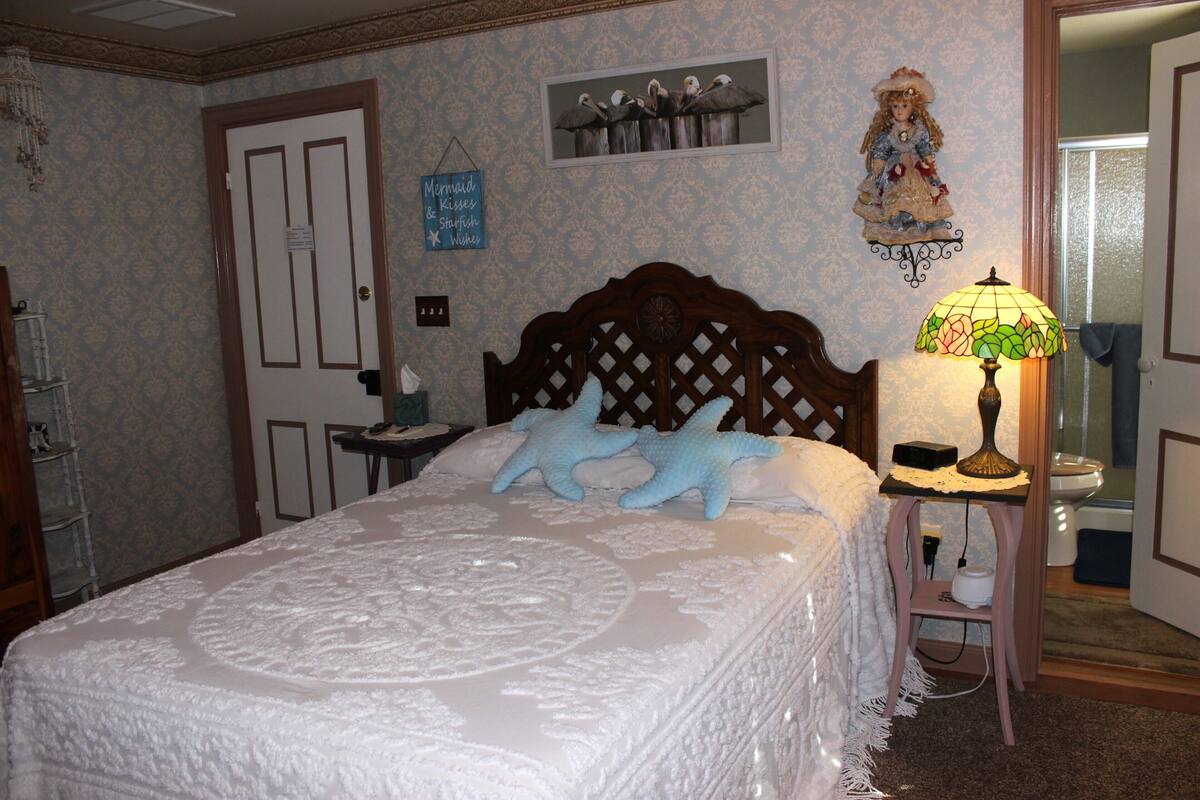 A bedroom with a large bed, a wooden headboard, a nightstand with a lamp, a dresser, and a door with a sign.