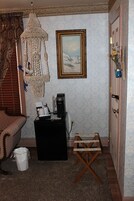 Una habitación con cama, cafetera, una mesita, una silla, un cubo de basura, una lámpara decorativa colgante y un cuadro en la pared.