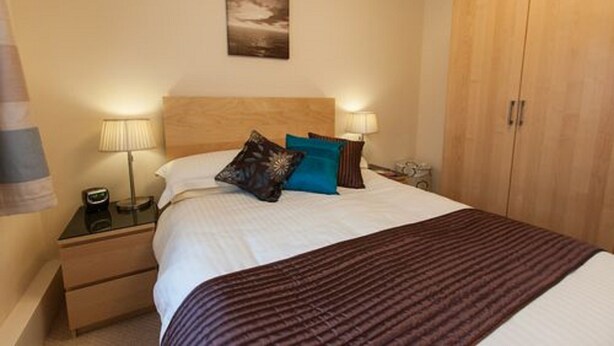 A hotel room with a bed, two bedside lamps, a nightstand, a wooden wardrobe, and a painting on the wall. - Cedars Apartment (Dunoon)