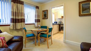 A living room with a brown leather sofa, a round table with two chairs, a kitchen visible through an open doorway, and a framed picture on the wall. - Cedars Apartment (Dunoon)