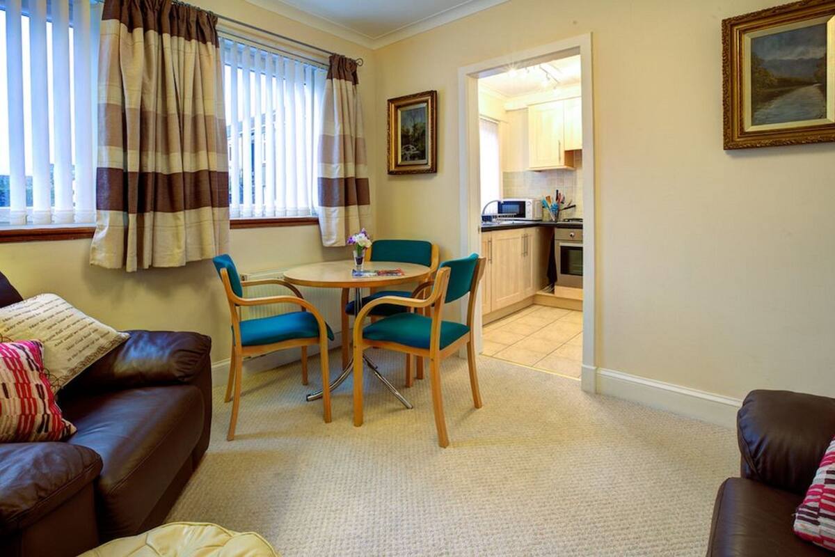 A living room with a brown leather sofa, a round table with two chairs, a kitchen visible through an open doorway, and a framed picture on the wall.