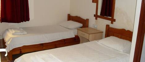 Quarto com duas camas de madeira, cada uma com colchão e almofadas brancas. Uma mesinha de cabeceira de madeira fica entre as camas, e uma pequena mesa com um abajur sobre ela.