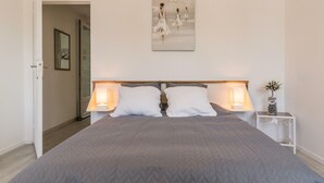 A bedroom with a large bed, two bedside lamps, a framed picture on the wall, and a small table with a plant.