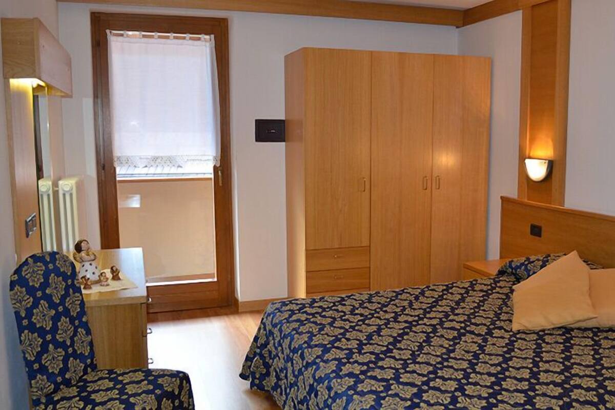 A bedroom with a bed, a chair, a wardrobe, a window with curtains, and a wall-mounted light fixture.