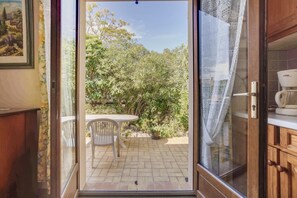Une porte coulissante ouverte donne sur un patio agrémenté d’une table et de chaises, le tout entouré de verdure.