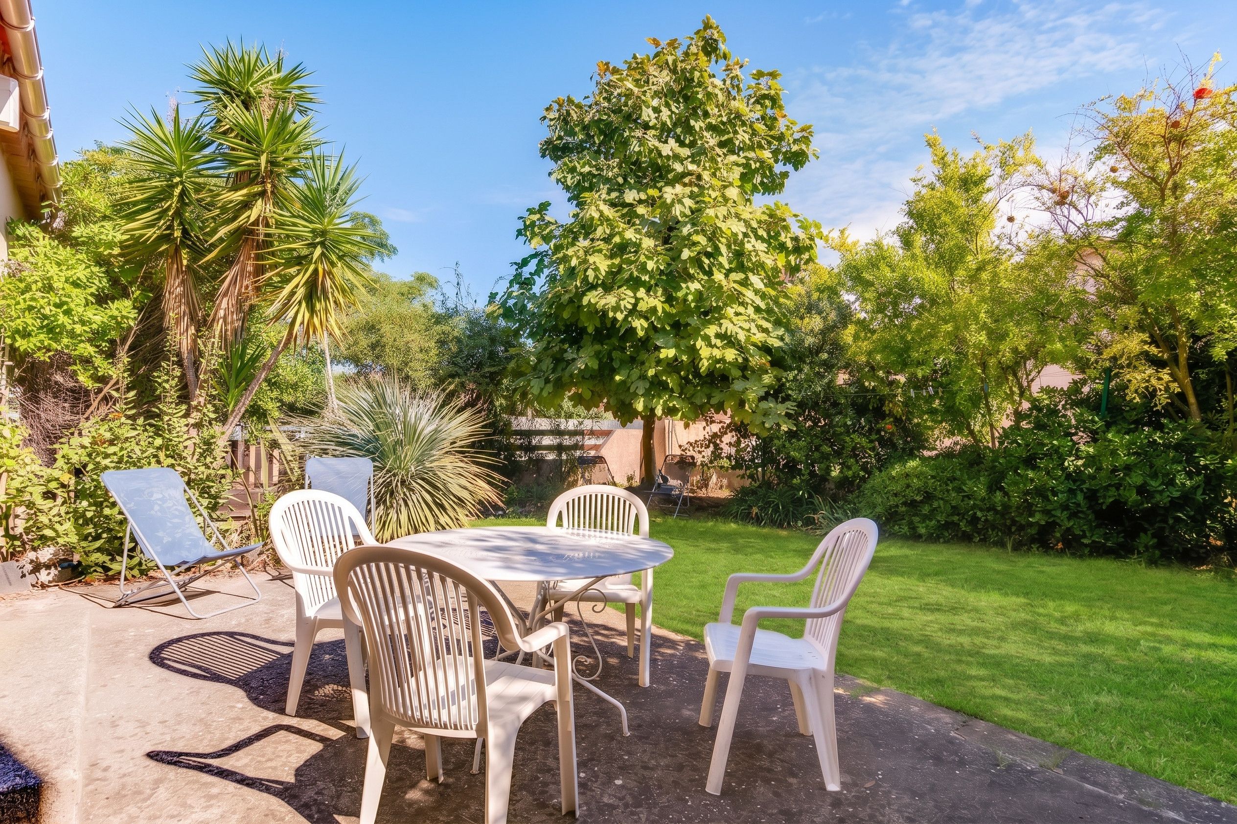 Eine Terrasse mit einem runden Tisch und vier Stühlen, umgeben von üppiger Bepflanzung und verschiedenen Bäumen.