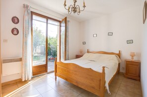 A bedroom with a wooden bed, a chandelier, a sliding glass door to a garden, and a small bedside table with a lamp. - Homerez - House in Agde (Agde)