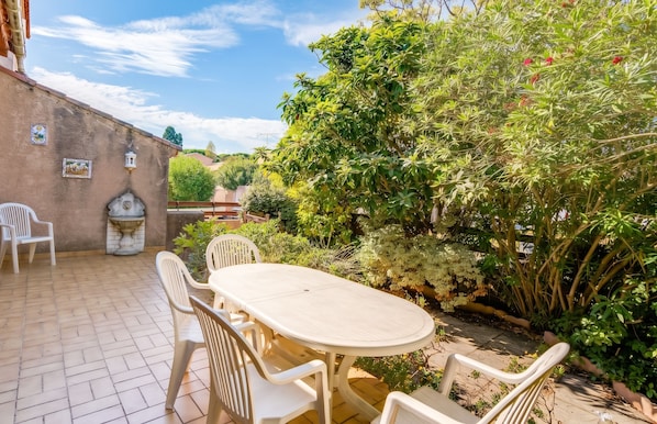 A patio with a round table and chairs, surrounded by lush greenery and a brick wall. - Homerez - House in Agde (Agde)