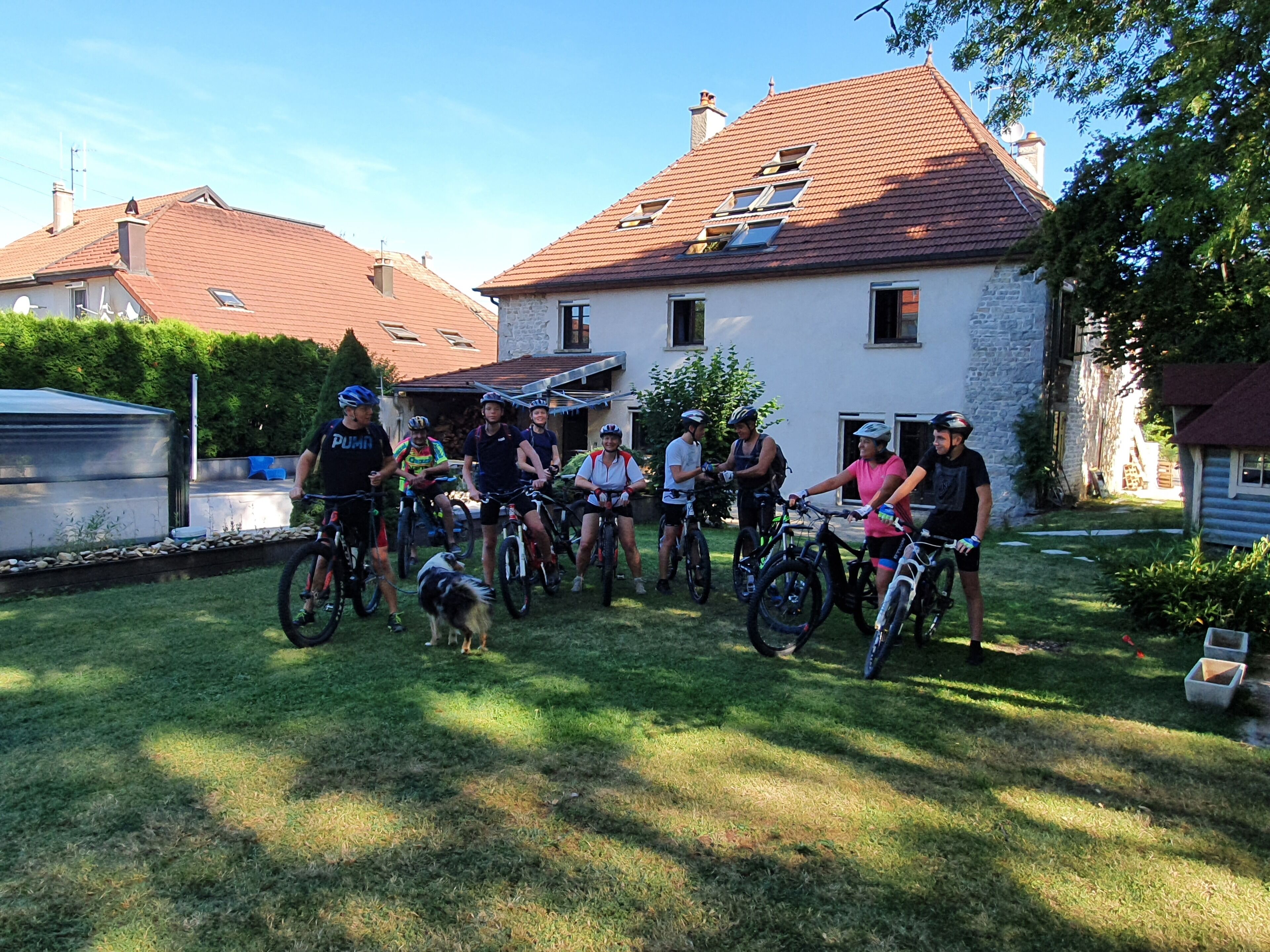 A group of cyclists gathered in a grassy area in front of a house with a stone facade and a tiled roof.