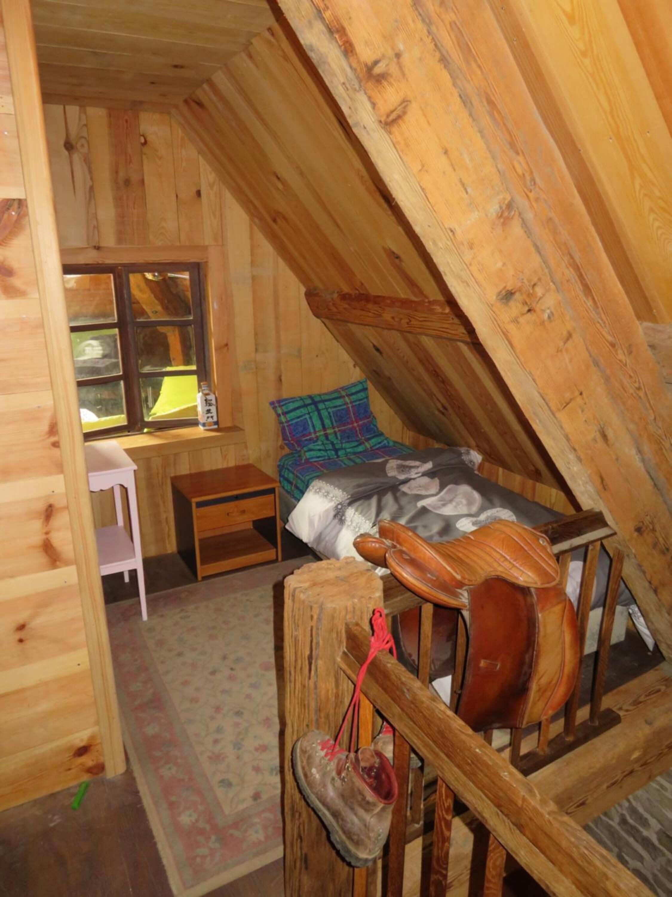A cozy attic bedroom with a wooden sloped ceiling, a bed with a plaid blanket, a small desk, and a chair.