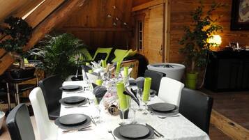 A dining area with a high ceiling and wooden beams, featuring a table set for six with chairs, a potted plant, and a painting on the wall.