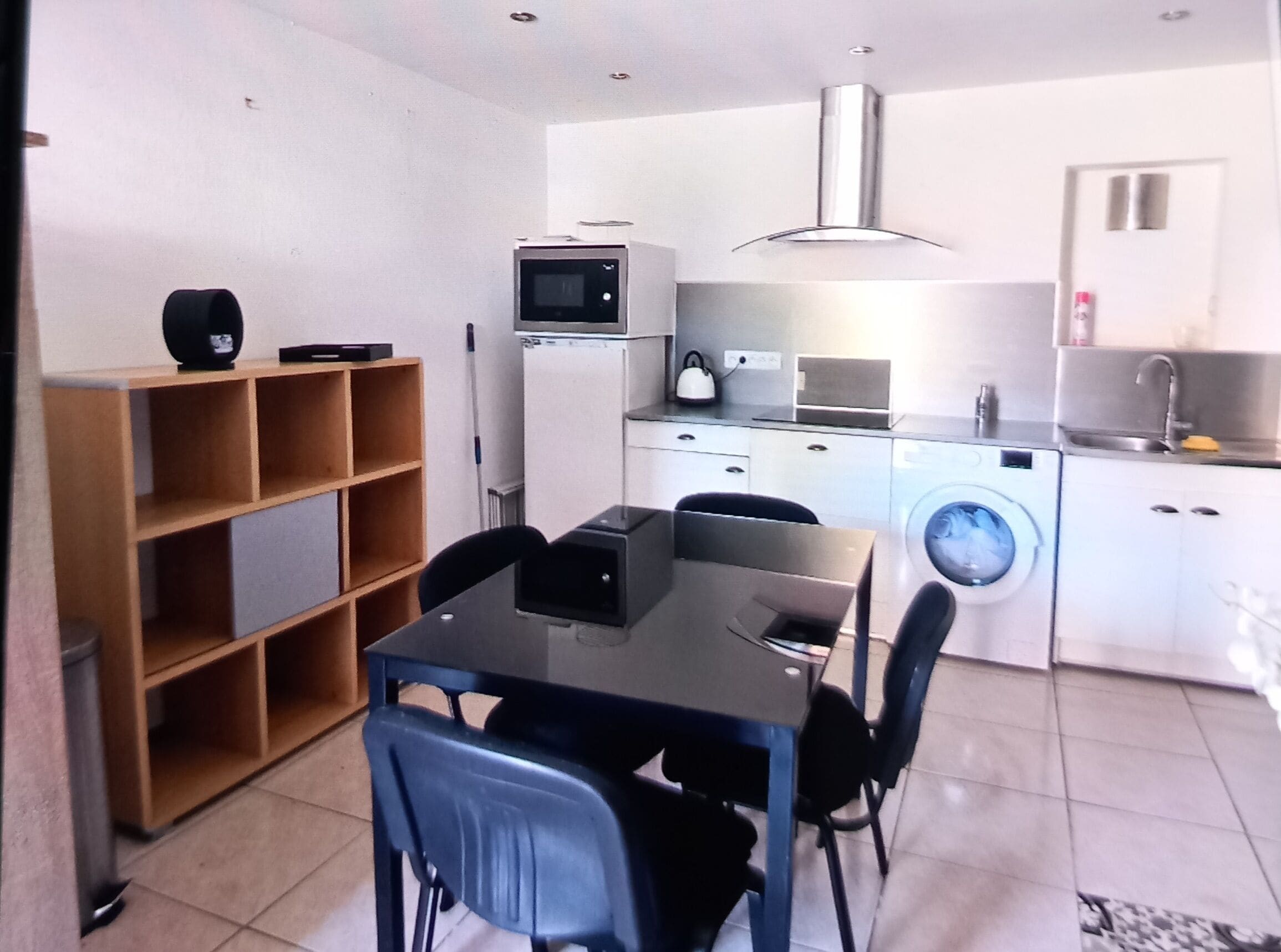 A compact kitchen with a microwave, sink, and washing machine. There is a dining table with chairs and a bookshelf.