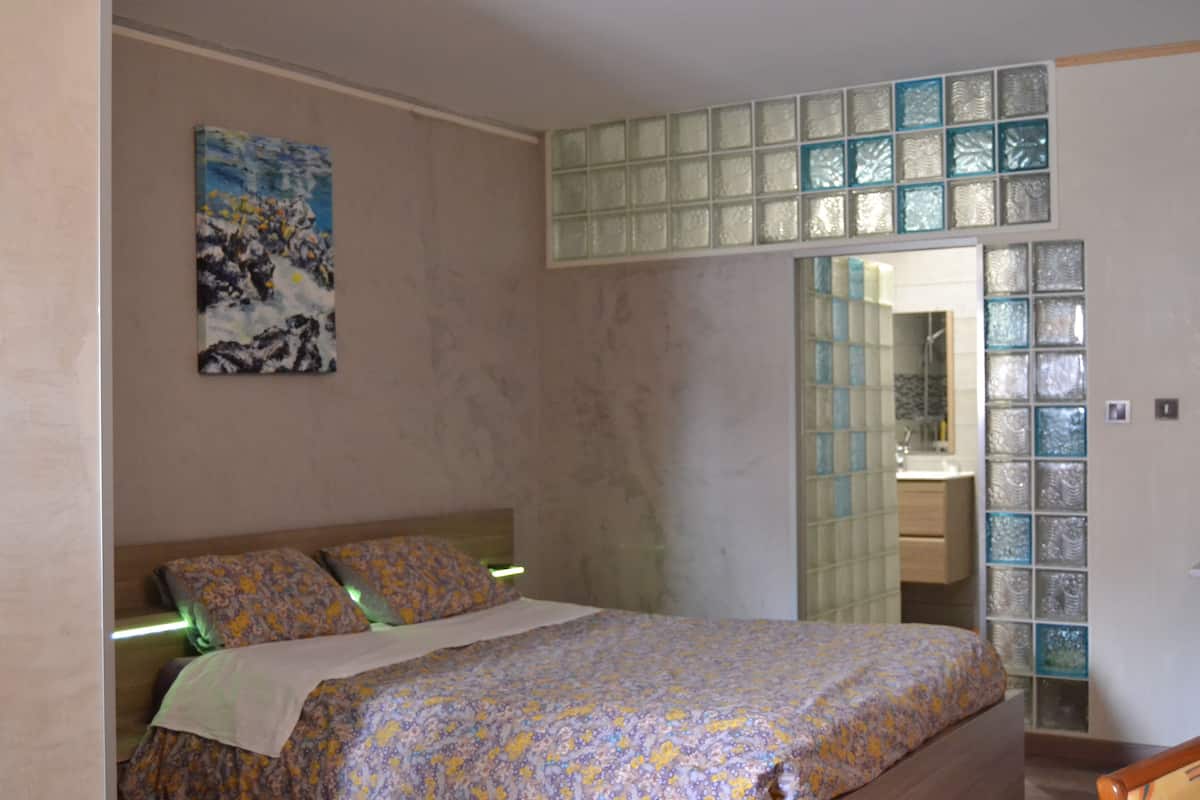 A bedroom with a bed, a painting on the wall, and a bathroom visible through a glass door.