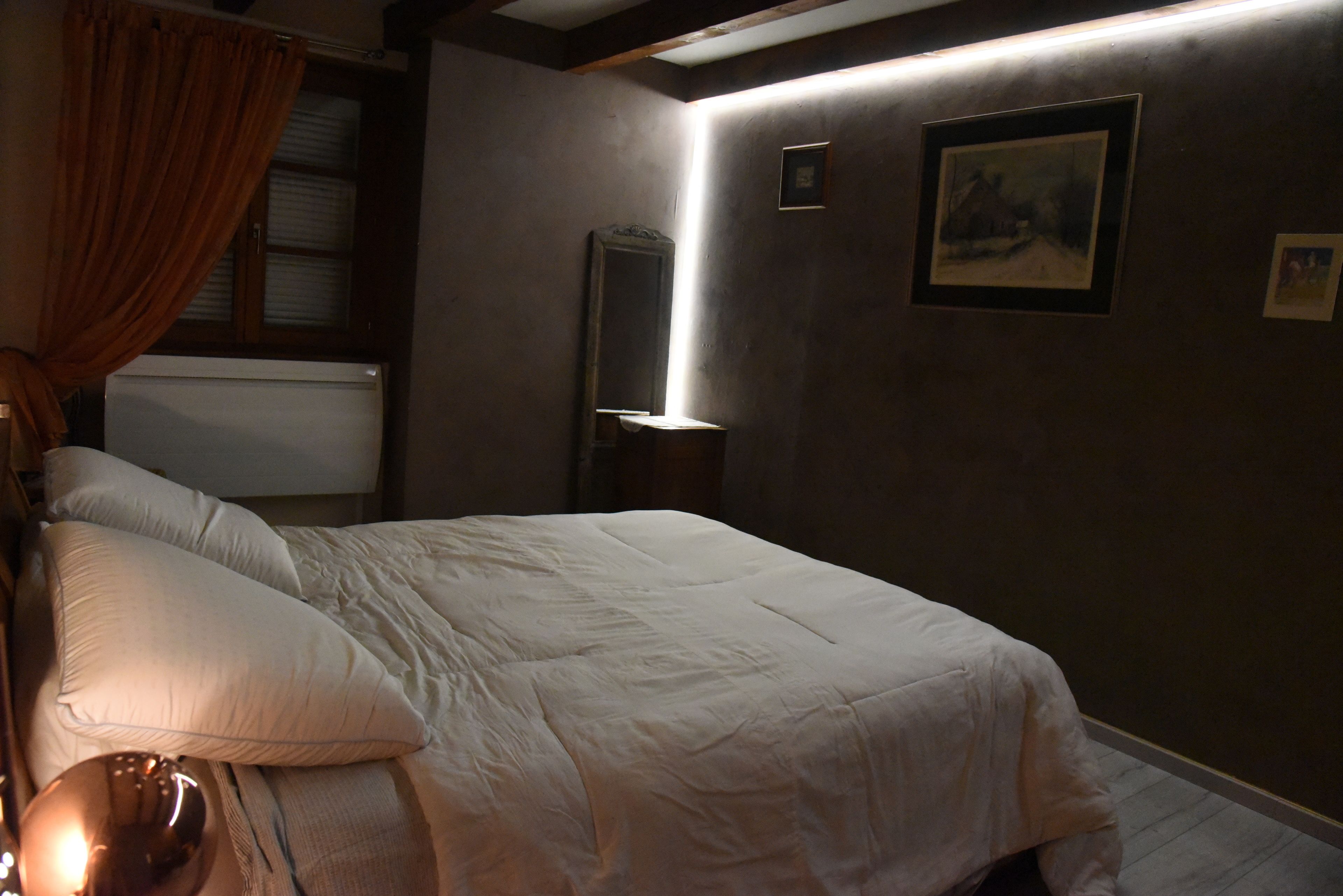 Une chambre à coucher avec un lit, une table de chevet, une lampe, une fenêtre avec des rideaux et des tableaux encadrés accrochés au mur.