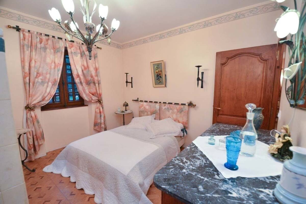 Quarto com cama, mesa de cabeceira, cômoda, lustre e janela com cortinas.