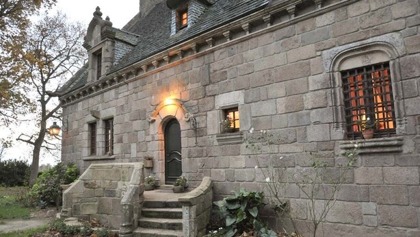 A stone building with a central entrance, a small porch with steps, and a window with a barred design.