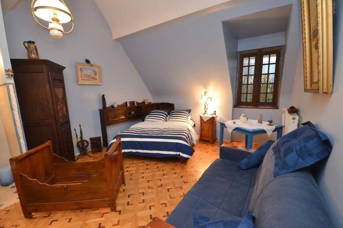 Um quarto com teto inclinado, uma cama de madeira, um sofá azul, uma mesinha com abajur e uma janela com vista.