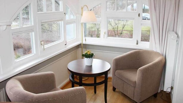 A room with two armchairs, a round table with a lamp, and large windows with curtains. - Café & Hotel Daheim (Masserberg)