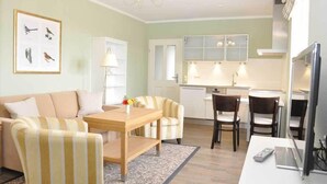 A modern living room with a sofa, armchairs, a coffee table, and a dining area with chairs. There is a kitchen in the background with cabinets and a refrigerator. - Café & Hotel Daheim (Masserberg)
