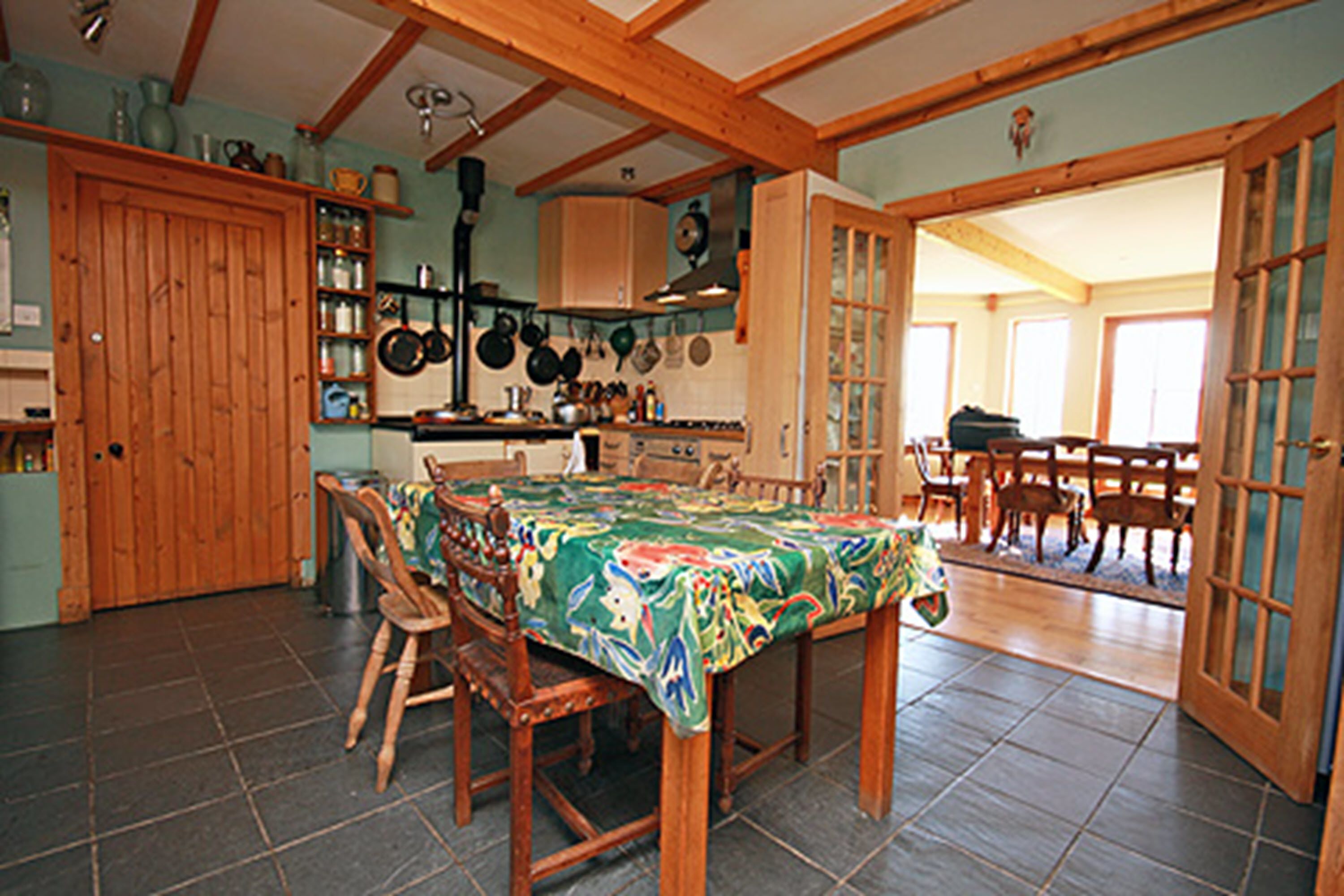 A dining area with a wooden table and chairs, a kitchen with hanging pots, and a door leading to another room.