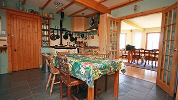 A dining area with a wooden table and chairs, a kitchen with hanging pots, and a door leading to another room.
