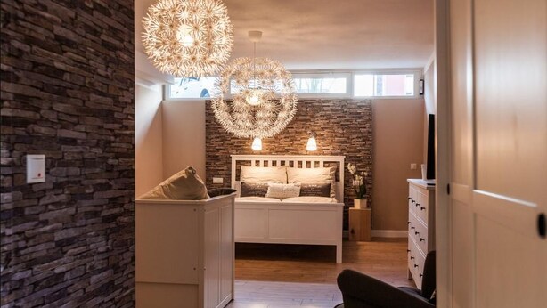 A bedroom with a stone wall, a bed with a headboard, bedside tables, and a large circular light fixture. - Haus am See B&B (Bornheim)