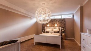 A bedroom with a white bed, a stone accent wall, a hanging light fixture, and a window with blinds. - Haus am See B&B (Bornheim)