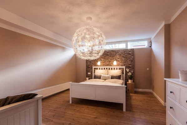 A bedroom with a white bed, a stone accent wall, a hanging light fixture, and a window with blinds.