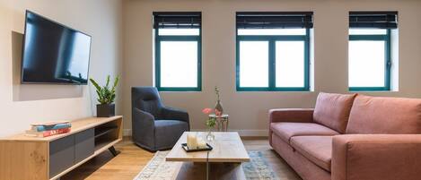 A modern living room with a flat-screen TV, a grey armchair, a pink sofa, a wooden coffee table, and a small side table with a vase and candles.
