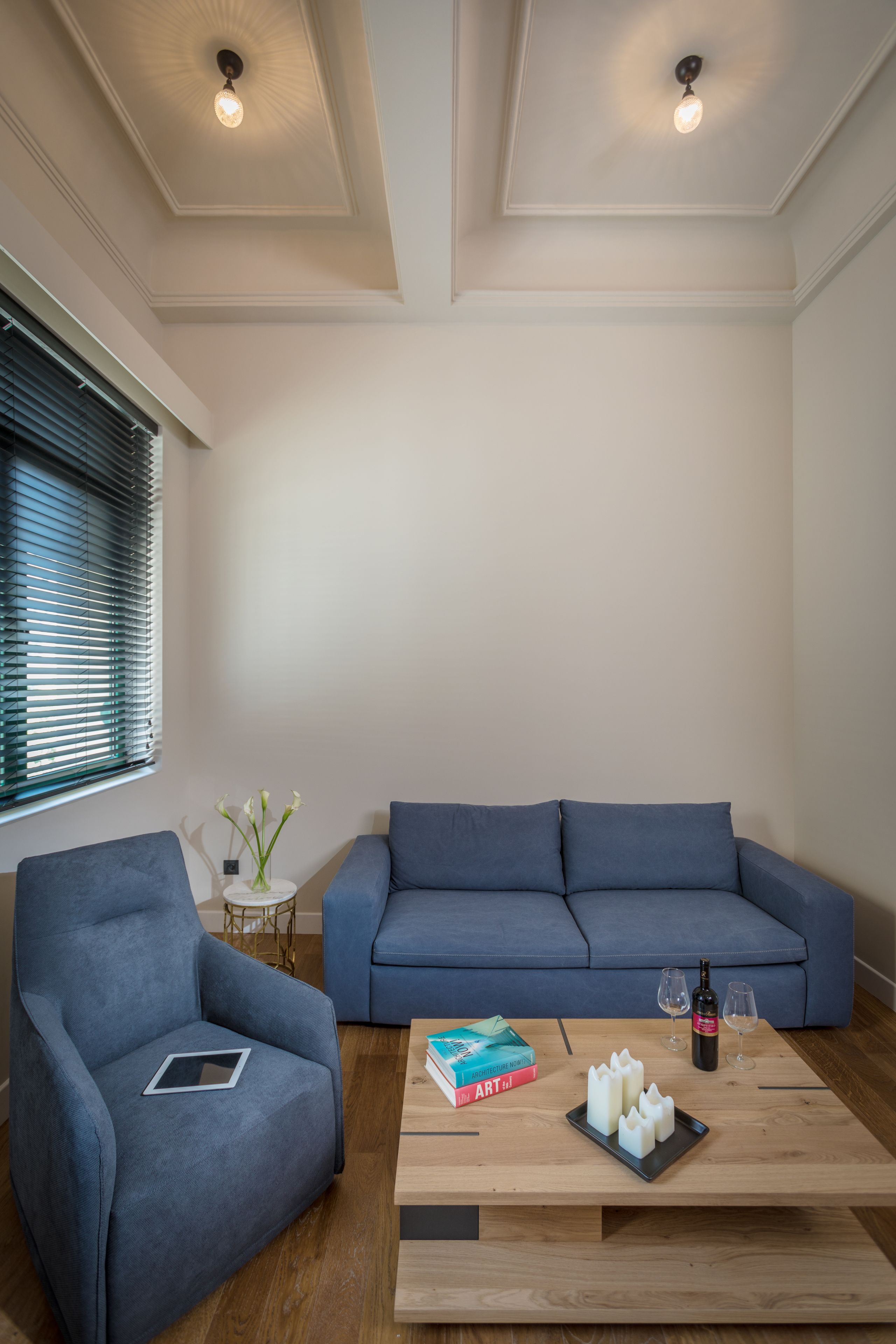 A modern living room with a blue sofa, a blue armchair, a wooden coffee table with books and candles, and a small side table with a vase and a bottle.