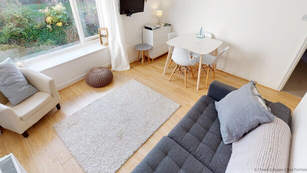 Una sala de estar acogedora con un sofá, una silla, una mesa de comedor con sillas, un televisor montado en la pared y un ventanal con vistas a la vegetación.