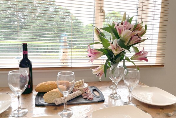 A dining table with a bottle of wine, a loaf of bread, a plate of cured meats, and a vase of flowers. - Windermere Lodge 2 Bedrooms (Windermere)