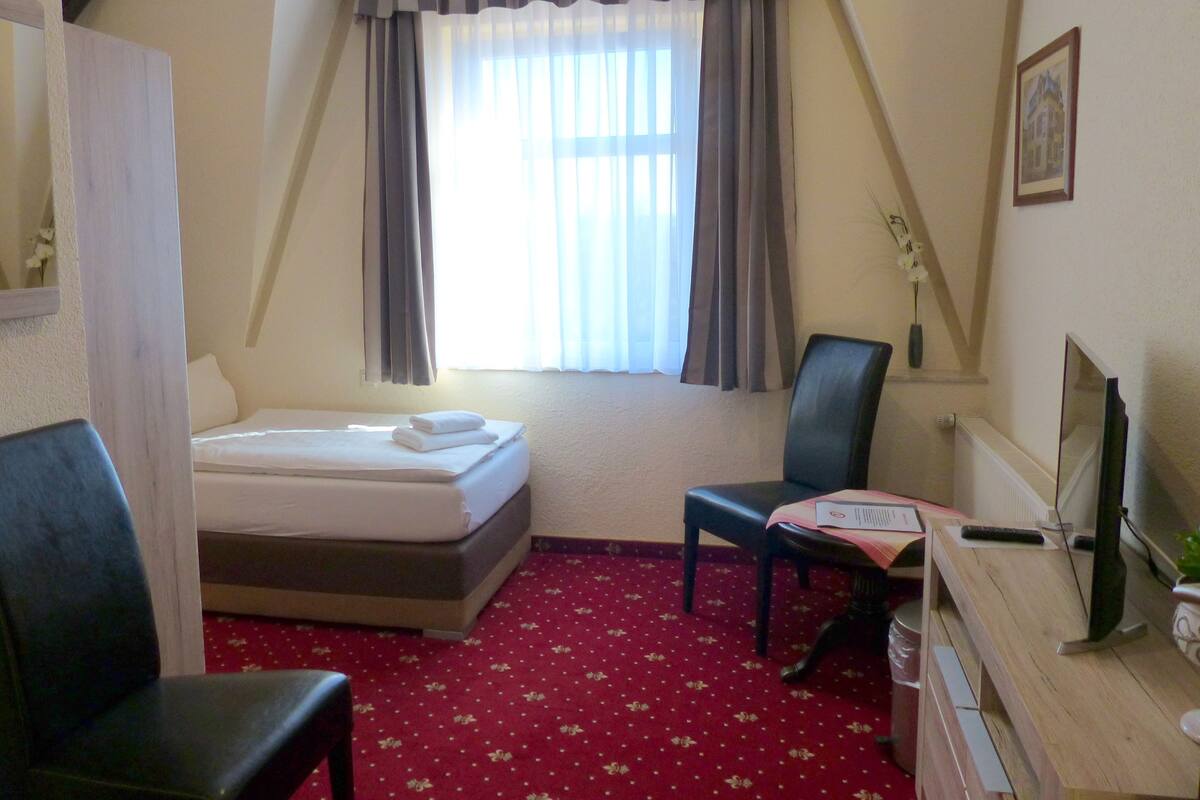 A hotel room with a bed, a desk with a computer, a chair, and a window with curtains.