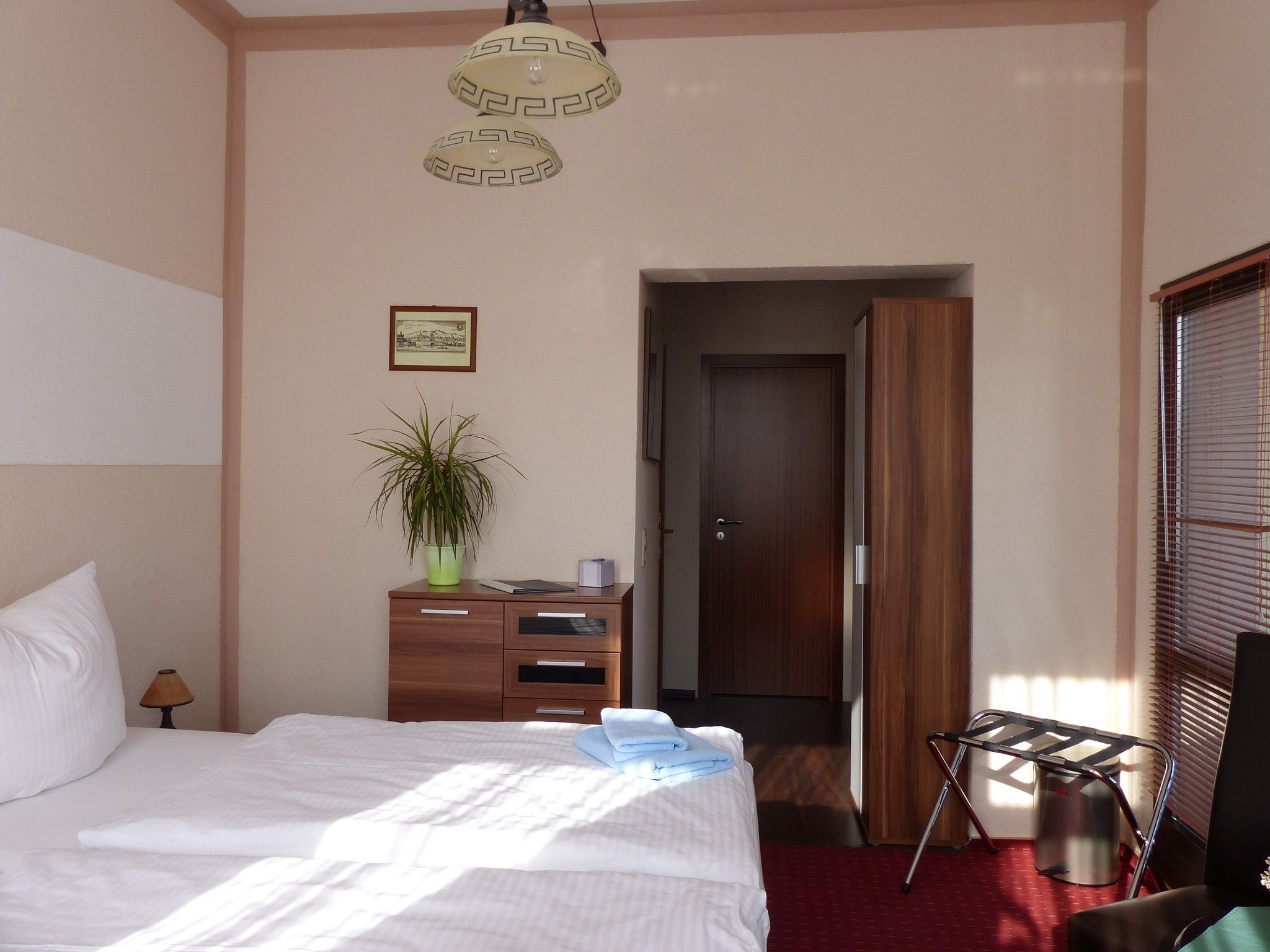 Een hotelkamer met een bed, een bureau met lades, een potplant, een deur en een raam met jaloezieën.