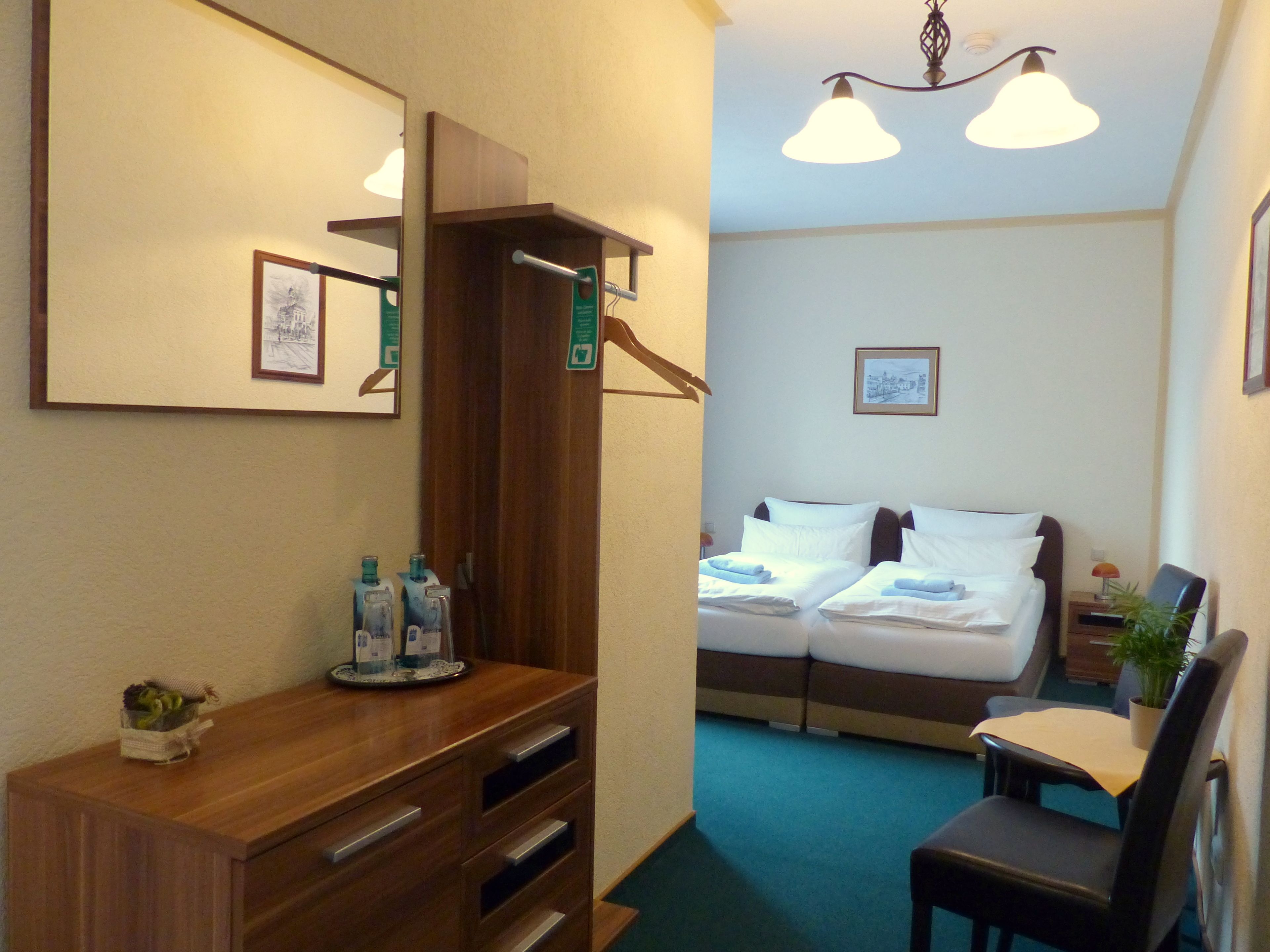 A hotel room with a wooden dresser, a desk, a chair, a bed with white linens, a small table with a plant, and a framed picture on the wall.