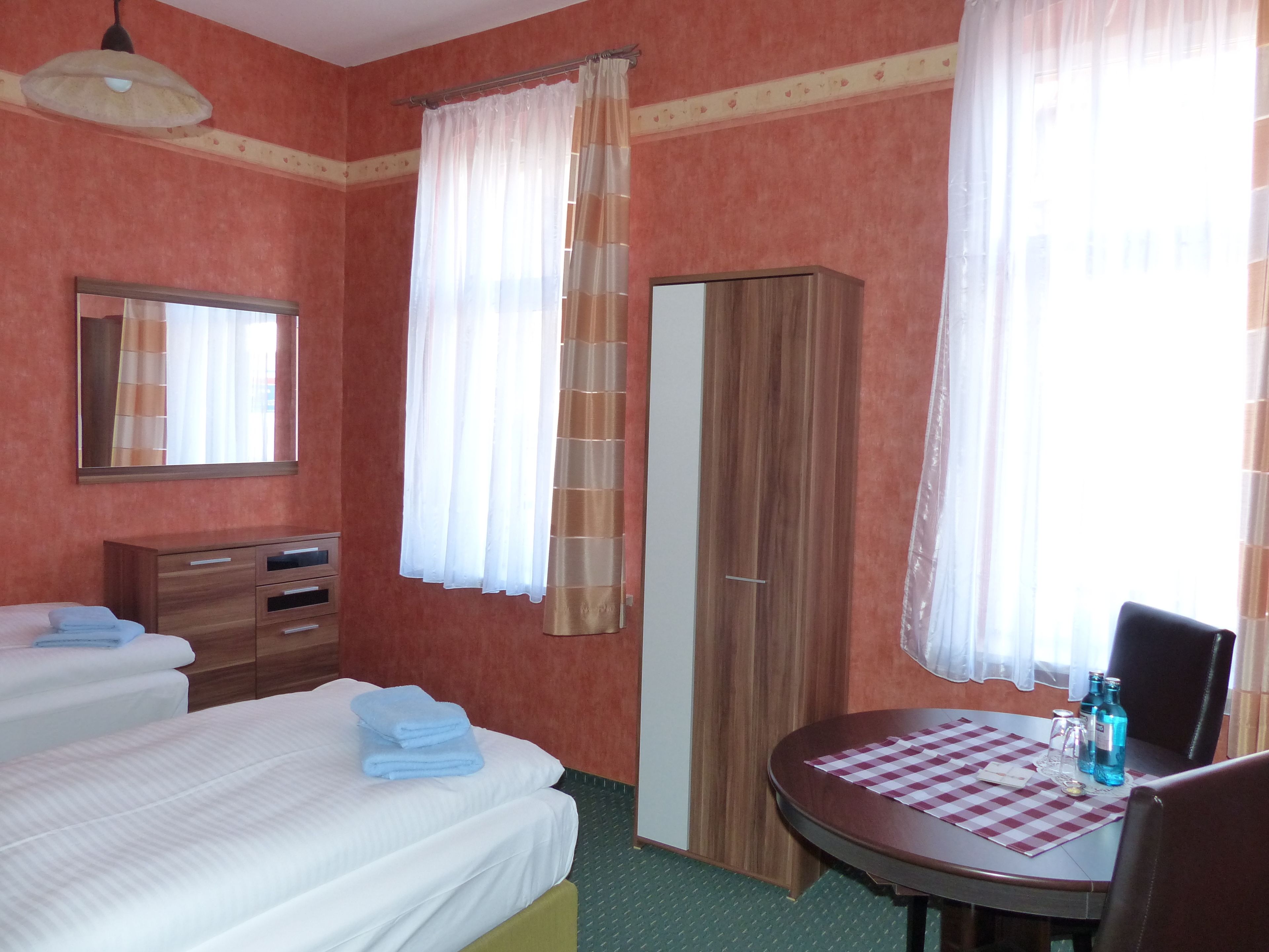 A hotel room with two beds, a wardrobe, a mirror, a table with a checkered tablecloth, and a window with curtains.